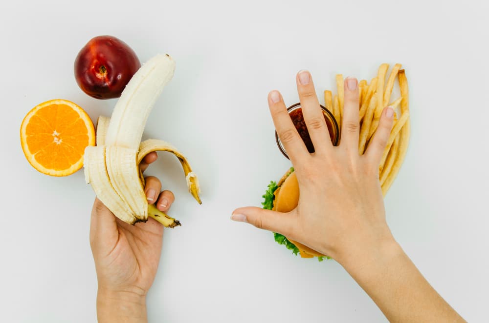 photo sur fond gris qui montre des fruits à gauche avec une main tenant une banane pelée, et des aliments gras, burger, sauce et frites avec une main posée dessus doigts écartés en signe de refus